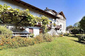 A stone building with a balcony, a wooden staircase, and a grassy yard. - Casa Alpinone: detached rustic house in the countryside with private garden and orchard (Crevoladossola)