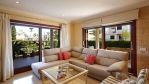 Ein Wohnzimmer mit einer Couch, Sesseln, einem Glastisch und großen Fenstern mit Vorhängen.