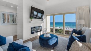 A modern living room with a large window offering a beach view, a flat-screen TV mounted on the wall, and a cozy seating area with blue upholstered furniture. - Gulf Front Condo w/Stunning Views, Private Beach Access, Gulf Front Pool! Tops'l Beach Resort (Miramar Beach)
