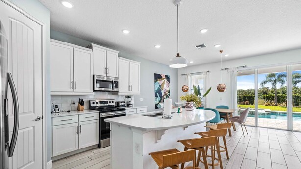 A modern kitchen with white cabinets, a central island with bar stools, and a view of a swimming pool through large windows. - Pool /Spa/Games Room/Movie Room/10 mins Disney/Restaurant and Bar/Modern /Chic (Kissimmee)