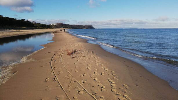 Uma extensa praia de areia com pegadas que levam à água, mar calmo e céu claro.