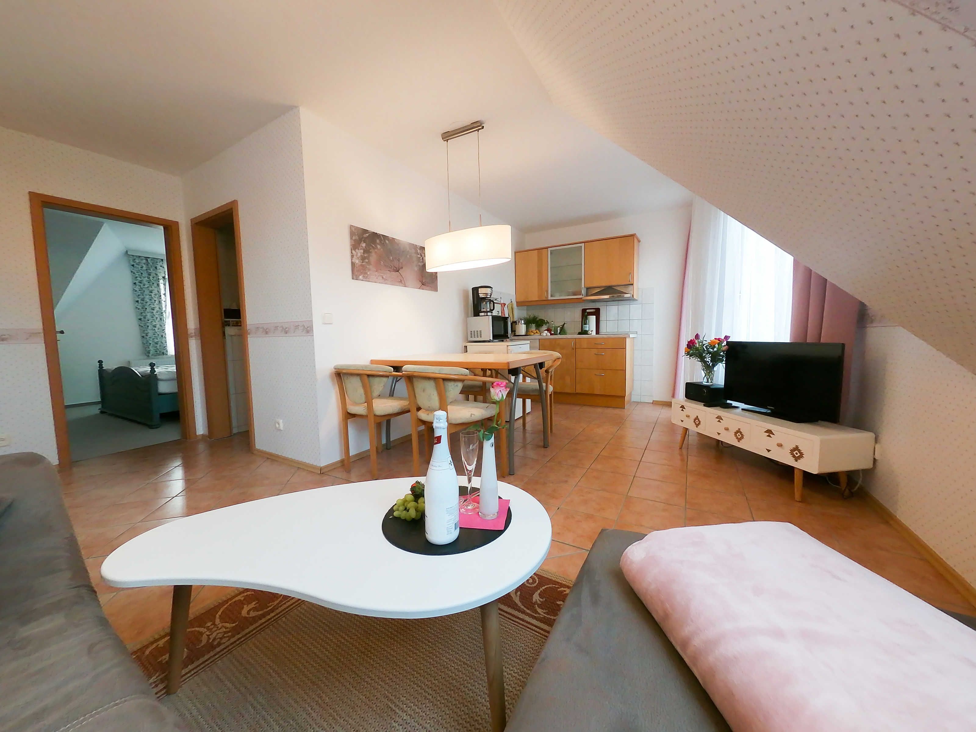 Ett modernt vardagsrum med ett vitt, böjt soffbord, en soffa och en tv som står på ett TV-bord. En matplats med trästolar och ett kök med träskåp och en mikrovågsugn syns också.