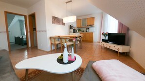 Uma sala de estar moderna com uma mesa de centro branca e curva, um sofá e uma televisão sobre um suporte. É visível uma área de jantar com cadeiras de madeira e uma cozinha com armários de madeira e micro-ondas.