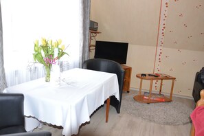 Una habitación con una mesa de comedor, una silla negra, una mesita redonda con un libro, un televisor sobre un mueble bajo y una pared con un diseño de corazón.