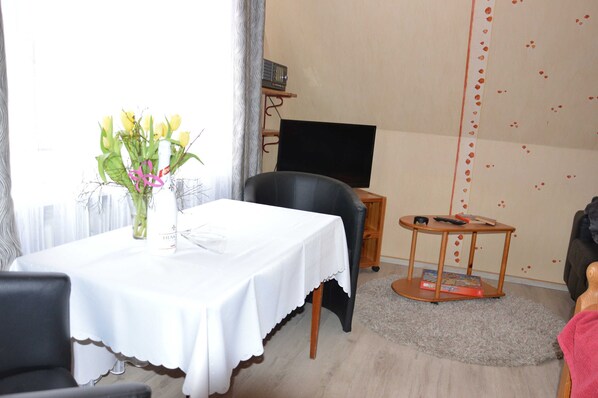 A room with a dining table, a black chair, a small round table with a book, a TV on a stand, and a wall with a heart pattern. - Muschelstrand 2-3 Pers - Fewo Vermietung Carmen Sagner (Baabe)