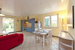 A compact kitchen with a microwave, refrigerator, and blue cabinets. There is a dining area with a white table and chairs, and a red sofa in the foreground.