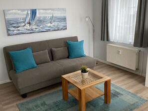 Uma sala de estar com um sofá, duas almofadas, uma mesa de centro com uma planta, um quadro de barcos à vela, um radiador e um abajur.