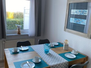 Uma área de jantar com uma mesa posta para dois, uma janela com cortinas, um radiador e um quadro na parede.