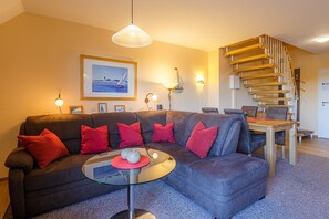 A modern living room with a grey sectional sofa, red cushions, a glass coffee table, a wooden spiral staircase, and a framed seascape painting on the wall. - Hafenmeisterei 8, Deutschland (Waren (Müritz))