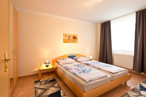 Una cama doble con ropa de cama floral, una mesita de noche con lámpara, una obra de arte en la pared y una ventana con cortinas.