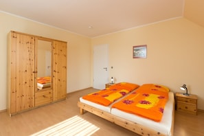 Une chambre avec une armoire en bois, un lit recouvert d’une couvre-lit à motifs floraux orange, une table de chevet avec une lampe et une porte.