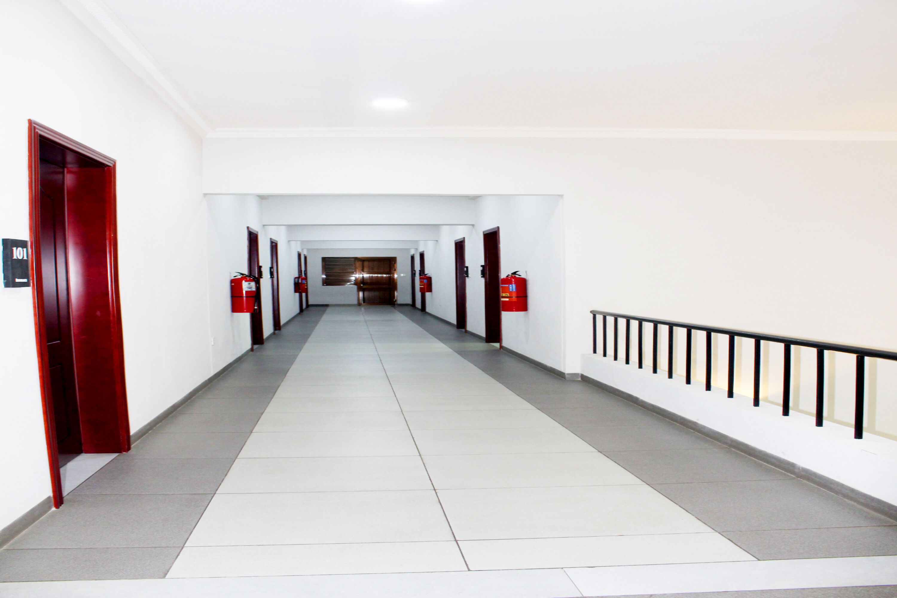 A long, narrow hallway with white walls and a tiled floor. There are doors on both sides, and a fire extinguisher is mounted on the wall.