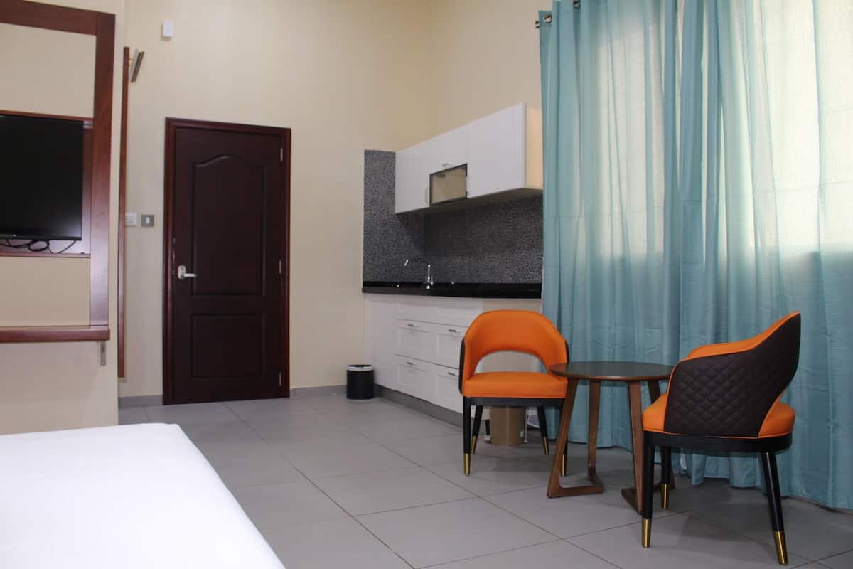 A small, well-lit room with a bed, a kitchenette, two orange chairs, and a blue curtain.