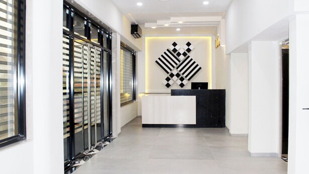 A modern reception area with a reception desk, a geometric wall art piece, and a glass door with blinds. - Ixoras Hotel (Kinshasa)