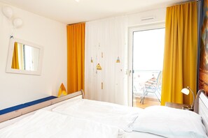 Quarto com cama, mesa de cabeceira com abajur, janela com cortinas e vista para a varanda.