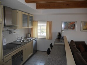 A compact kitchen with a stove, sink, and cabinets, a dining area with a table and chairs, and a window with curtains. - Kietzspeicher App. 21 (Waren (Müritz))