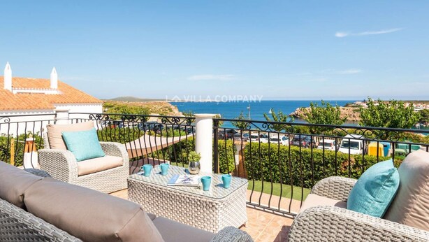 A balcony with wicker furniture, a view of the sea, and a building with a tiled roof. - Wonderful Villa Bellavista with Sea Views & Private Pool, Sleeps 8 (Arenal d'en Castell, Es Mercadal)