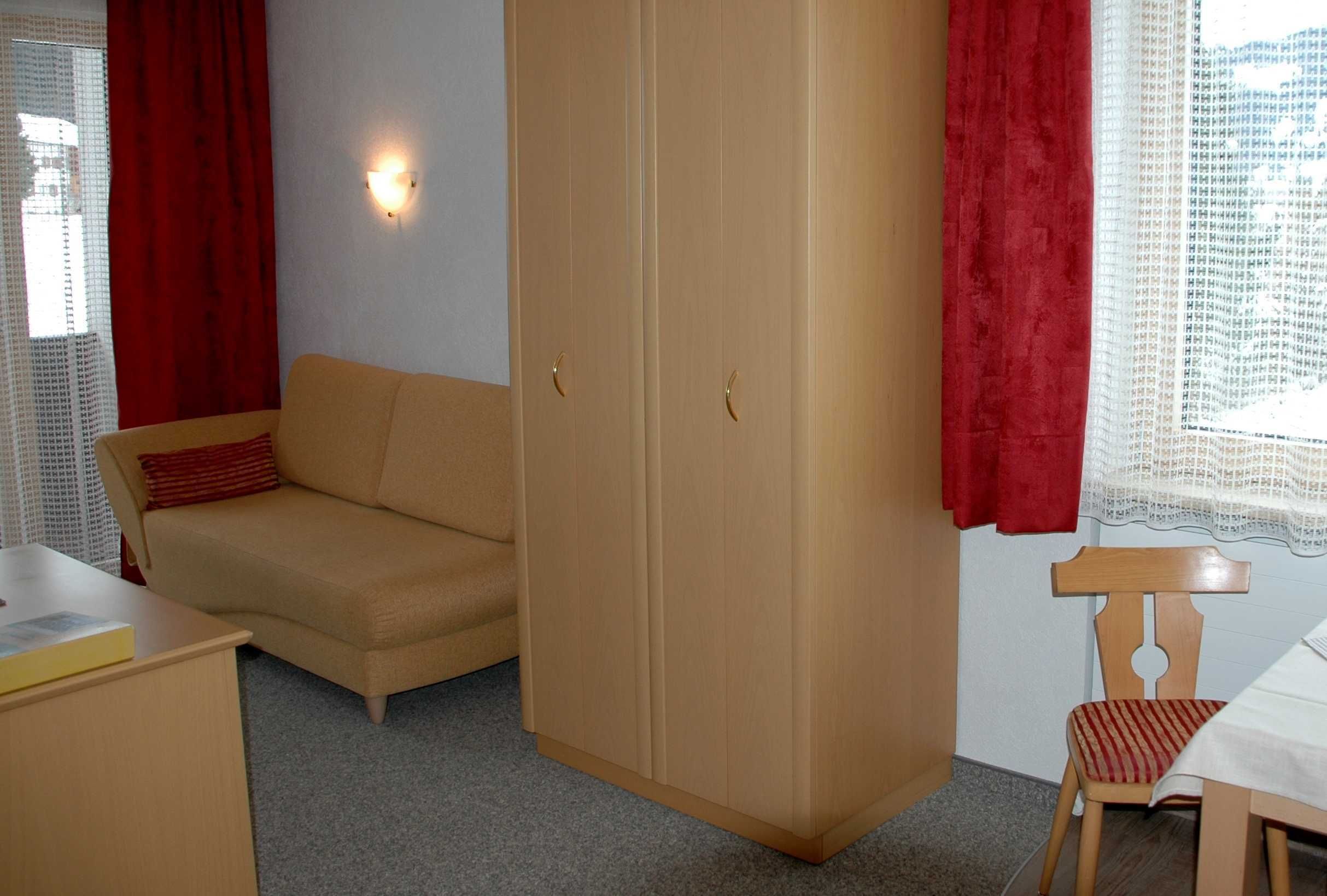 A room with a beige sofa, a wooden wardrobe, a small table, and a chair.