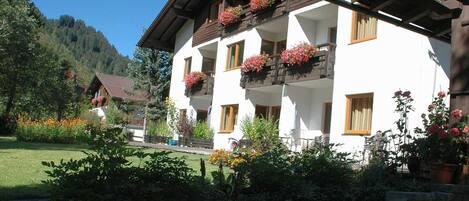 A white building with balconies, surrounded by greenery and a mountain backdrop.