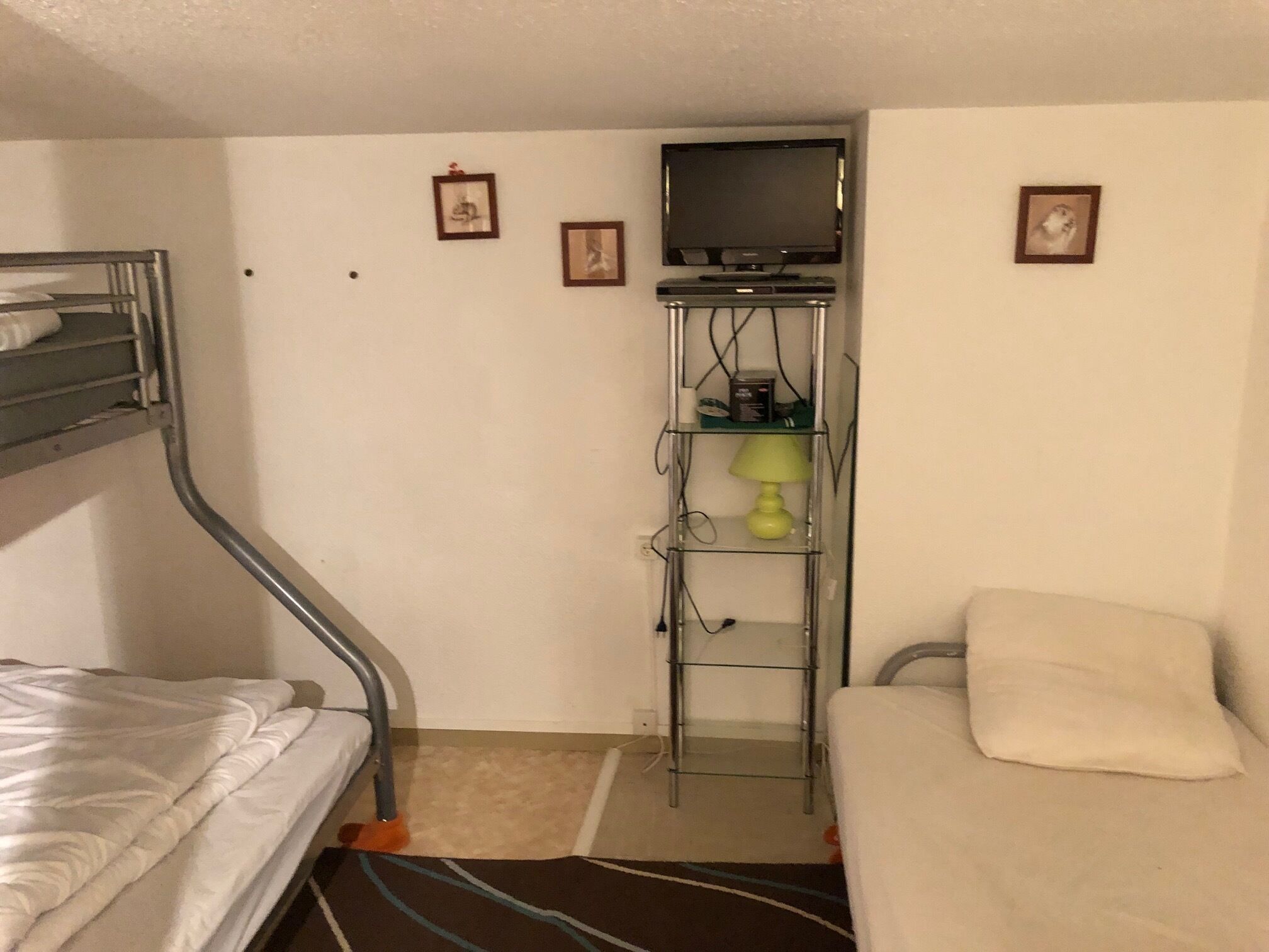 A compact hotel room with a bunk bed, a desk with a computer, a television, and a small shelf with a lamp.