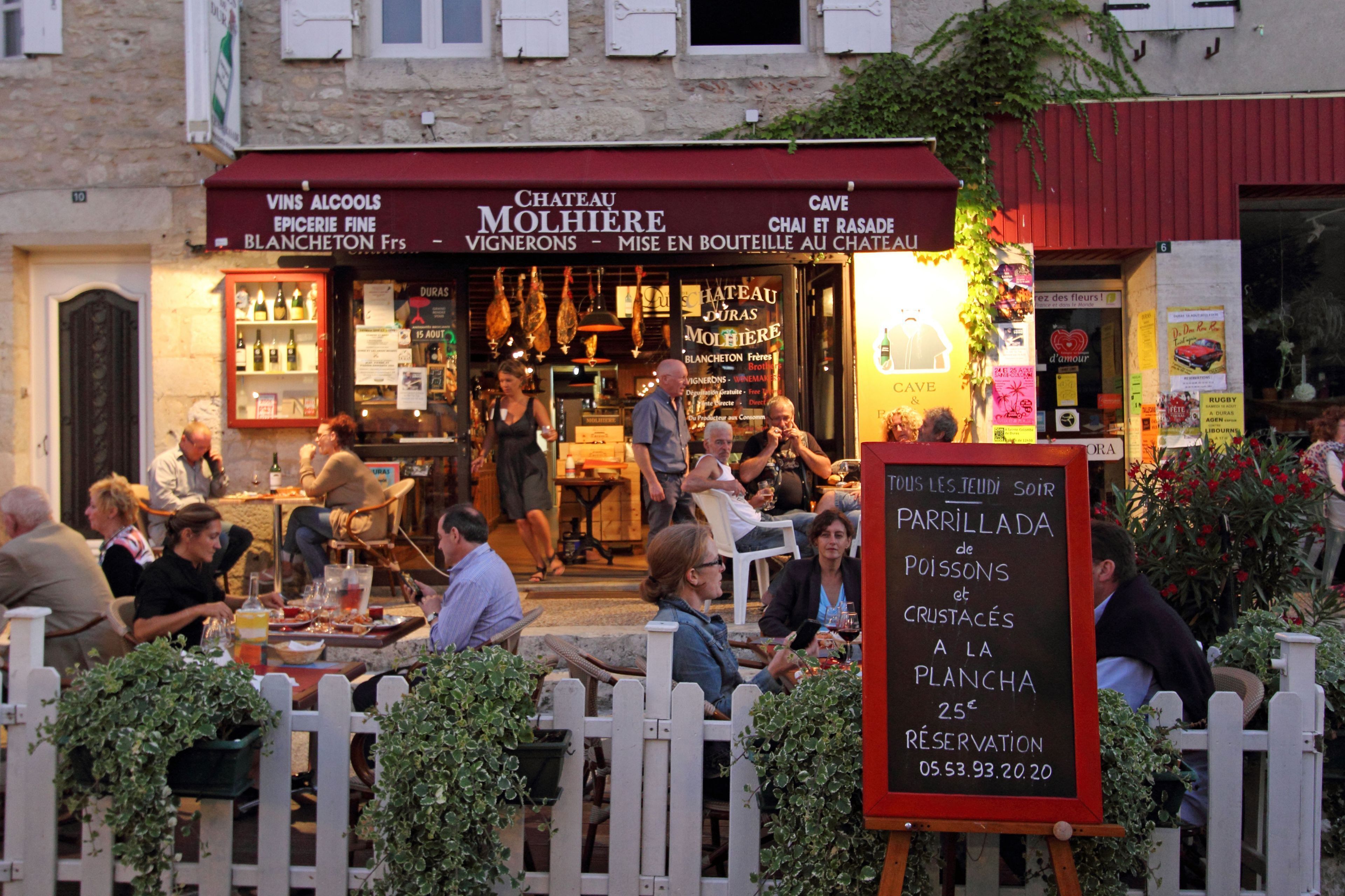 Un restaurante con terraza, un cartel que anuncia 'Parrillada de Poissons et Crustacés', y un toldo rojo con la inscripción 'Chateau Molhieres'.