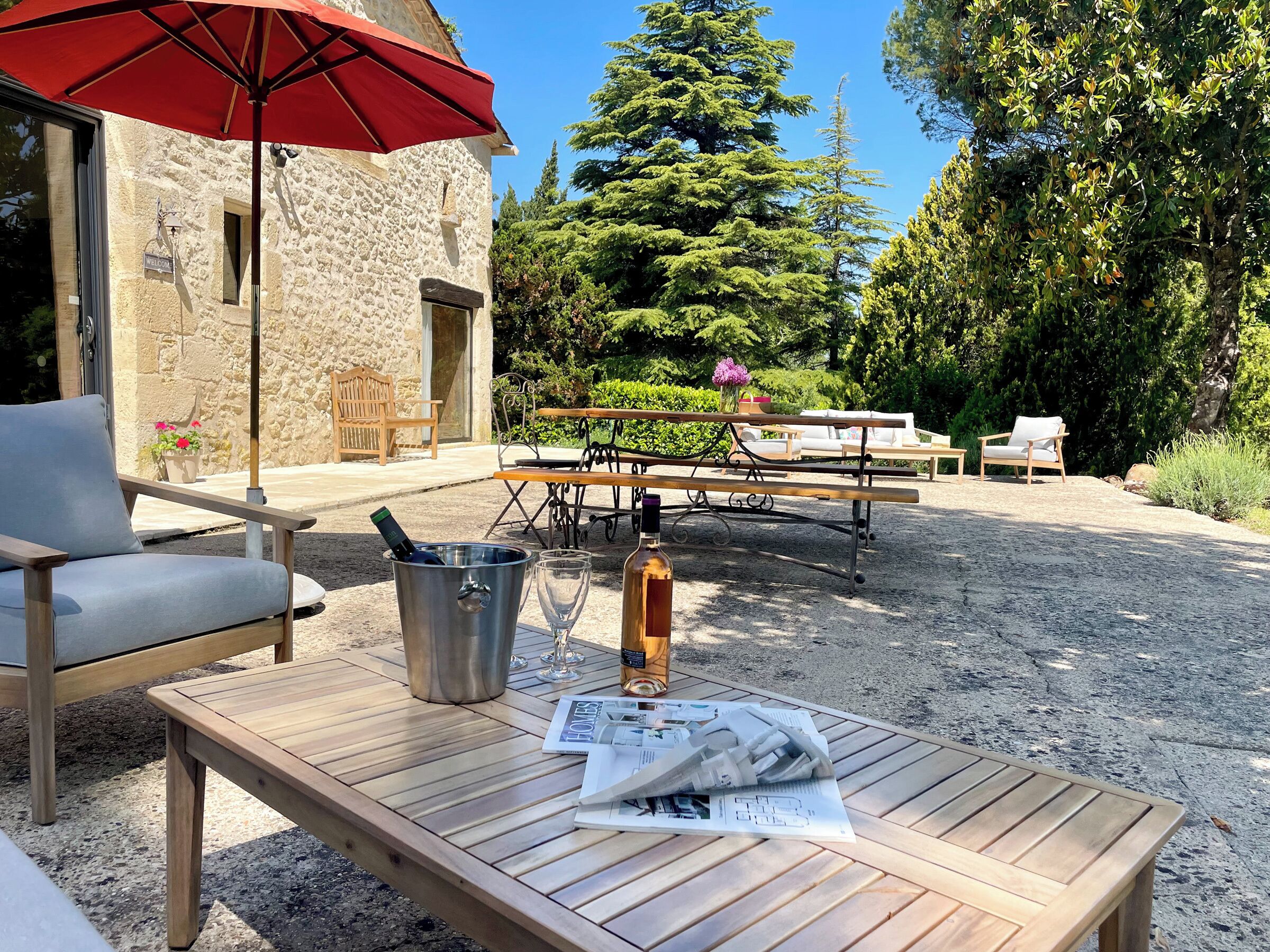 A wooden table with a bottle and glasses, a red umbrella, and outdoor seating.