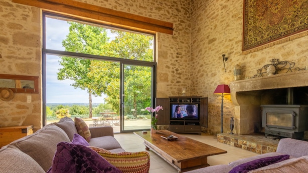 A cozy living room with a stone wall, large windows, a fireplace, and a television.