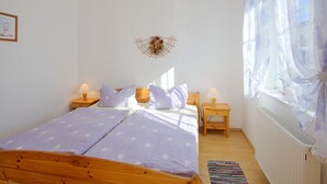 Quarto com cama de madeira, duas mesas de cabeceira com luminárias, radiador e janela com cortinas finas.