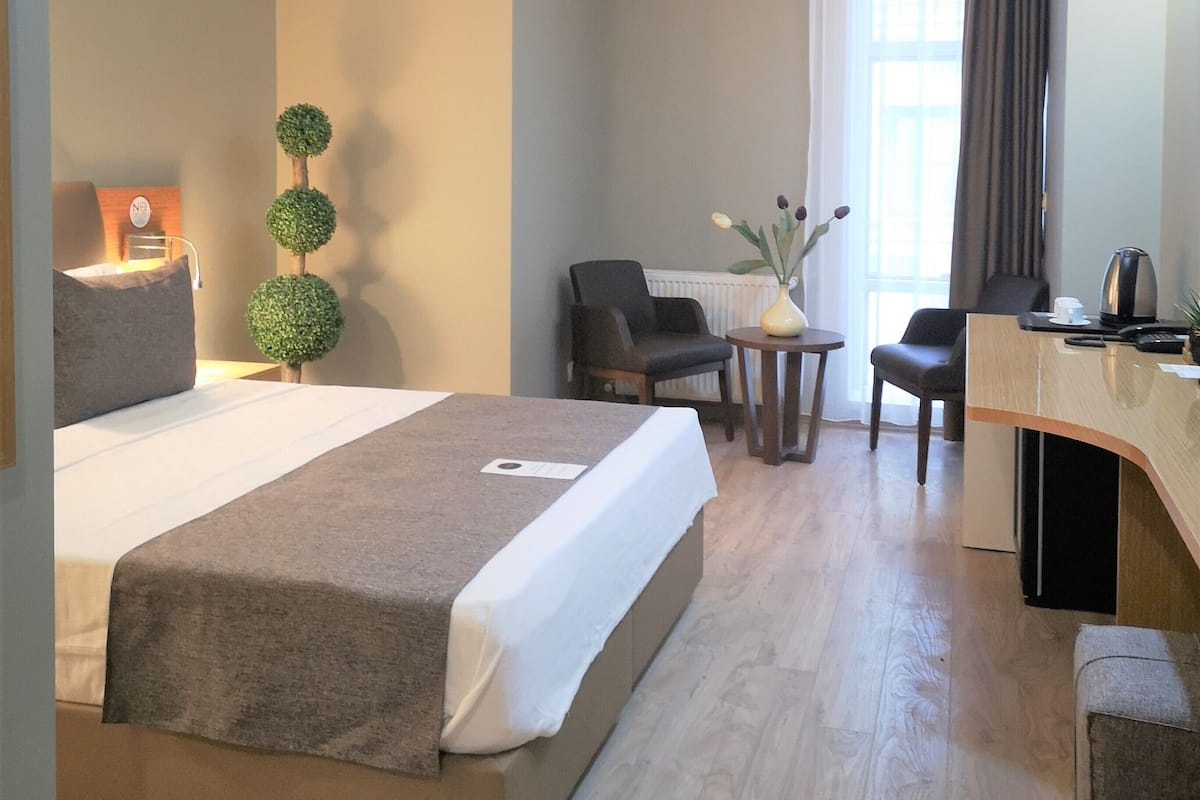 A hotel room with a bed, two chairs, a desk, a small table, and a vase with flowers.