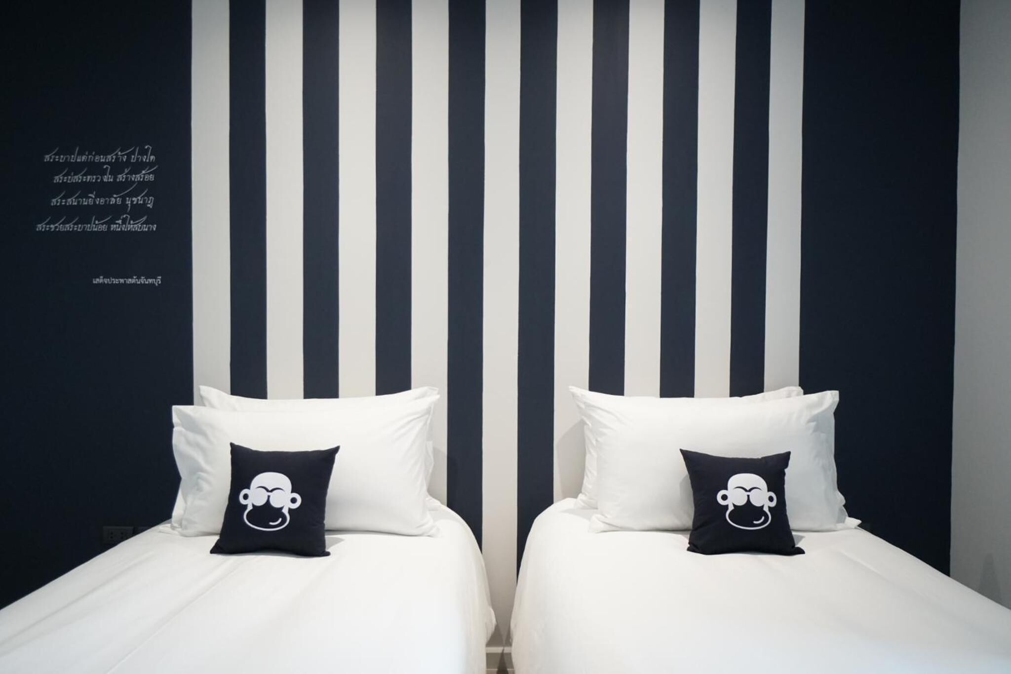 A hotel room with two beds, each with white bedding and black pillows featuring a cartoon character. The room has a striped wall design and a quote in a foreign language.
