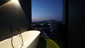 Un baño moderno con una bañera independiente, ducha y vistas a un paisaje urbano al atardecer.
