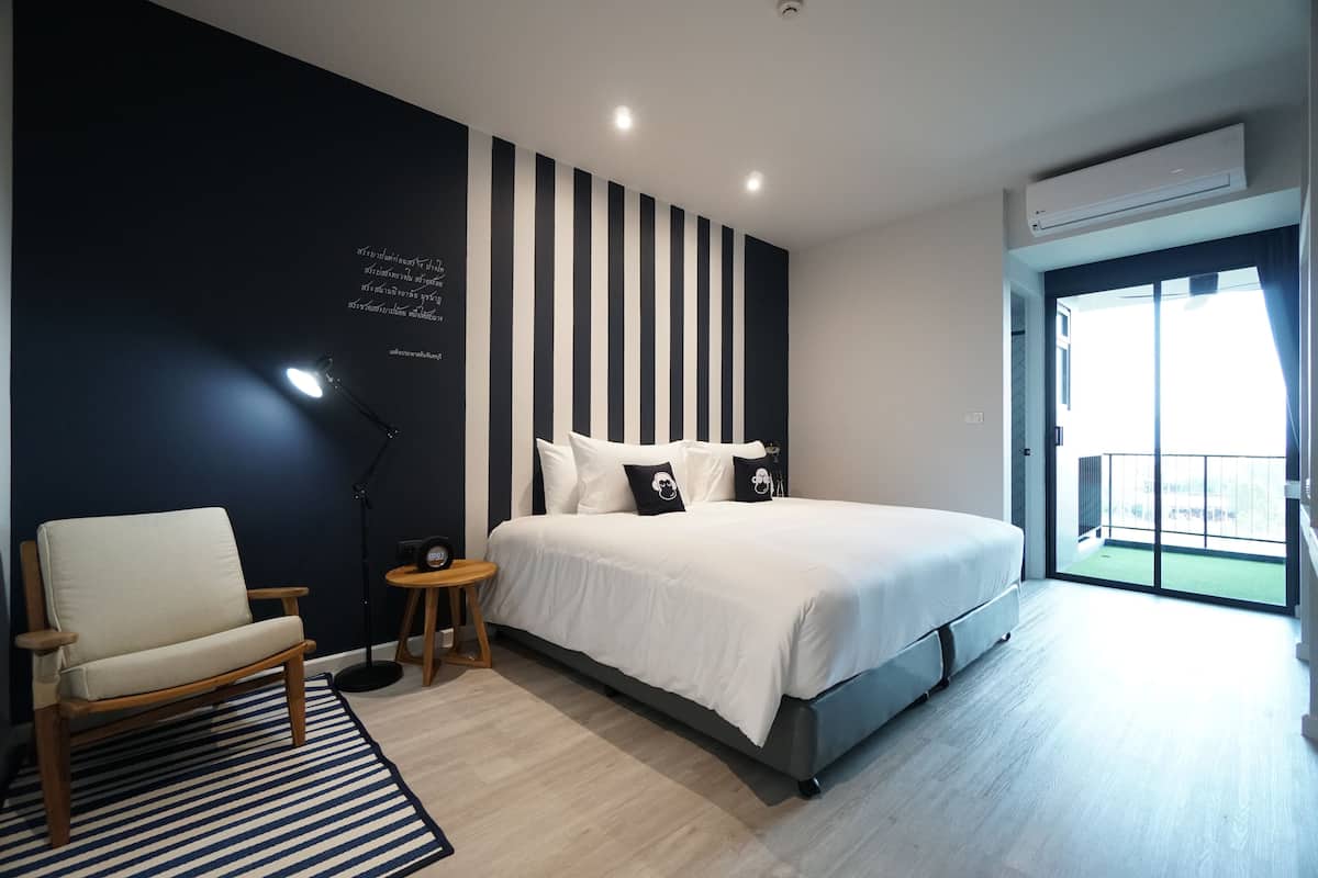 Ein modernes Schlafzimmer mit einem großen Bett, einem Nachttisch, einem Sessel, einer Stehlampe und Blick ins Freie vom Balkon aus.