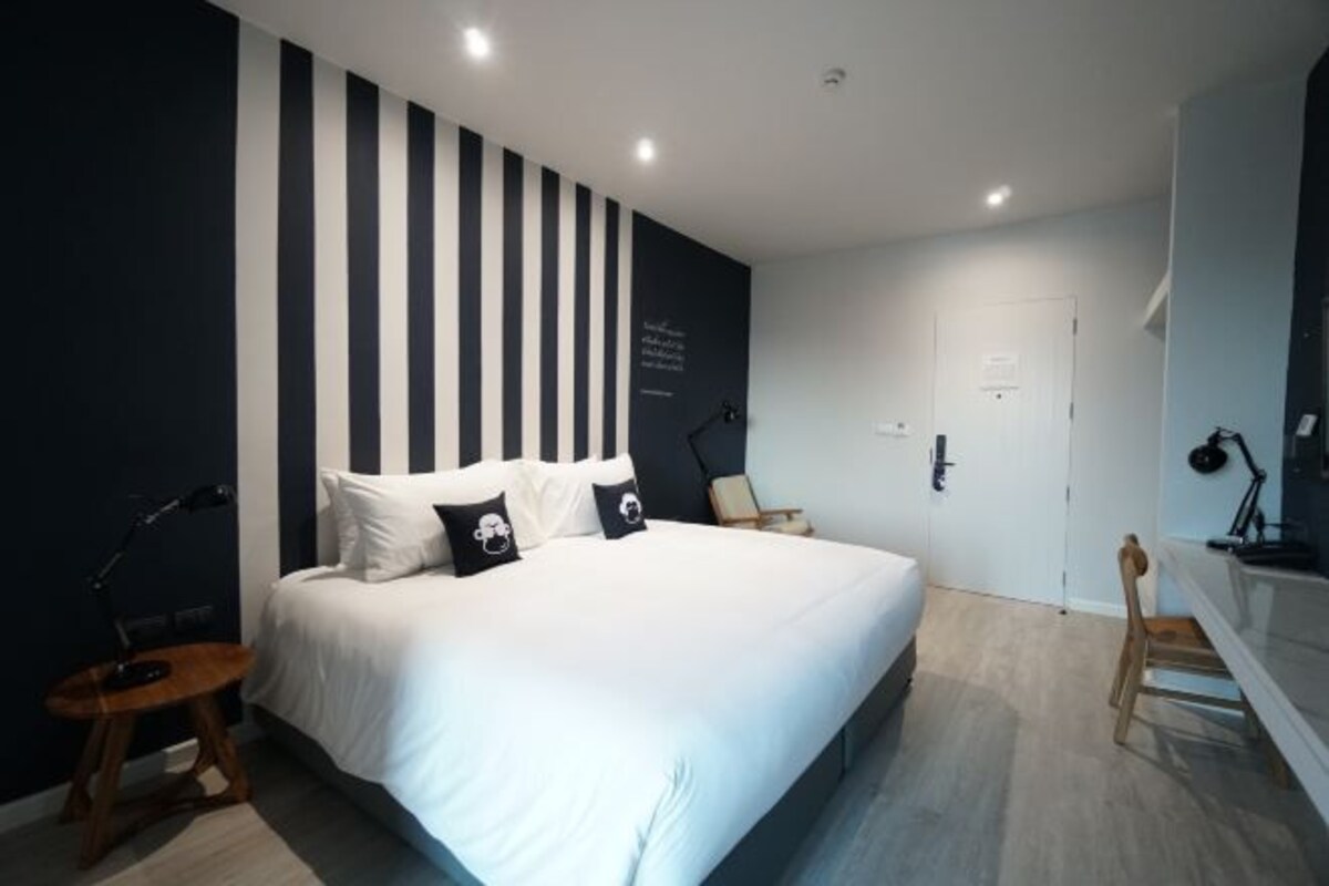 A modern bedroom with a large bed, a dark wall with vertical stripes, a bedside table with a lamp, and a wooden chair.