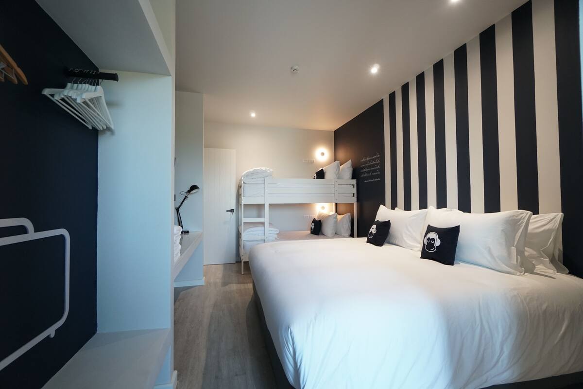 Ein modernes Hotelzimmer mit einem Etagenbett, einem großen Bett mit weißer Bettwäsche, einer schwarz-weiß gestreiften Wand und einem kleinen Schreibtisch mit einer Lampe.