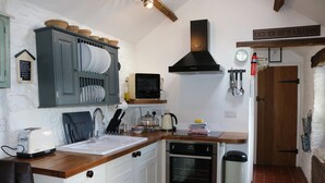 A compact kitchen with a built-in oven, a microwave, and a sink. There is a wooden countertop, a cabinet with dishes, and a sign on the wall. - Keepers Cottage - Cosy Peak District Retreat (Derbyshire)