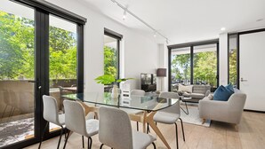 Ein modernes Wohnzimmer mit einem Glastisch, grauen Stühlen, einem grauen Sofa und einem großen Fenster mit Blick auf Bäume.
