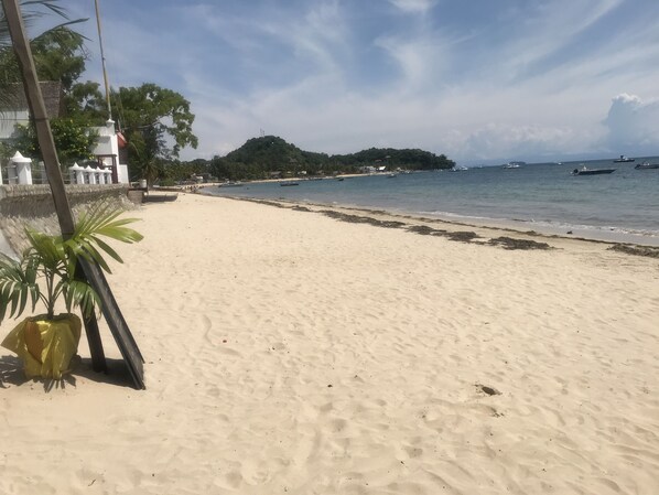 A sandy beach with a clear sky, a few boats in the water, and a distant hill.