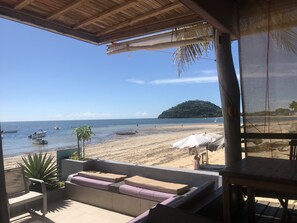 Vista a la playa desde un área de estar al aire libre techada, con cojines y una mesa.