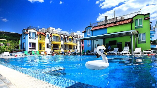 Une piscine avec un flotteur en forme de cygne, entourée de bâtiments aux couleurs vives et sous un ciel bleu clair.