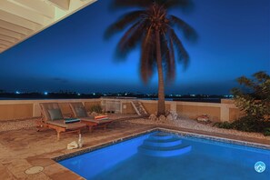 Poolbereich mit Liegestühlen und einer Palme, umgeben von einer Steinterrasse und Blick auf die Stadt bei Nacht.
