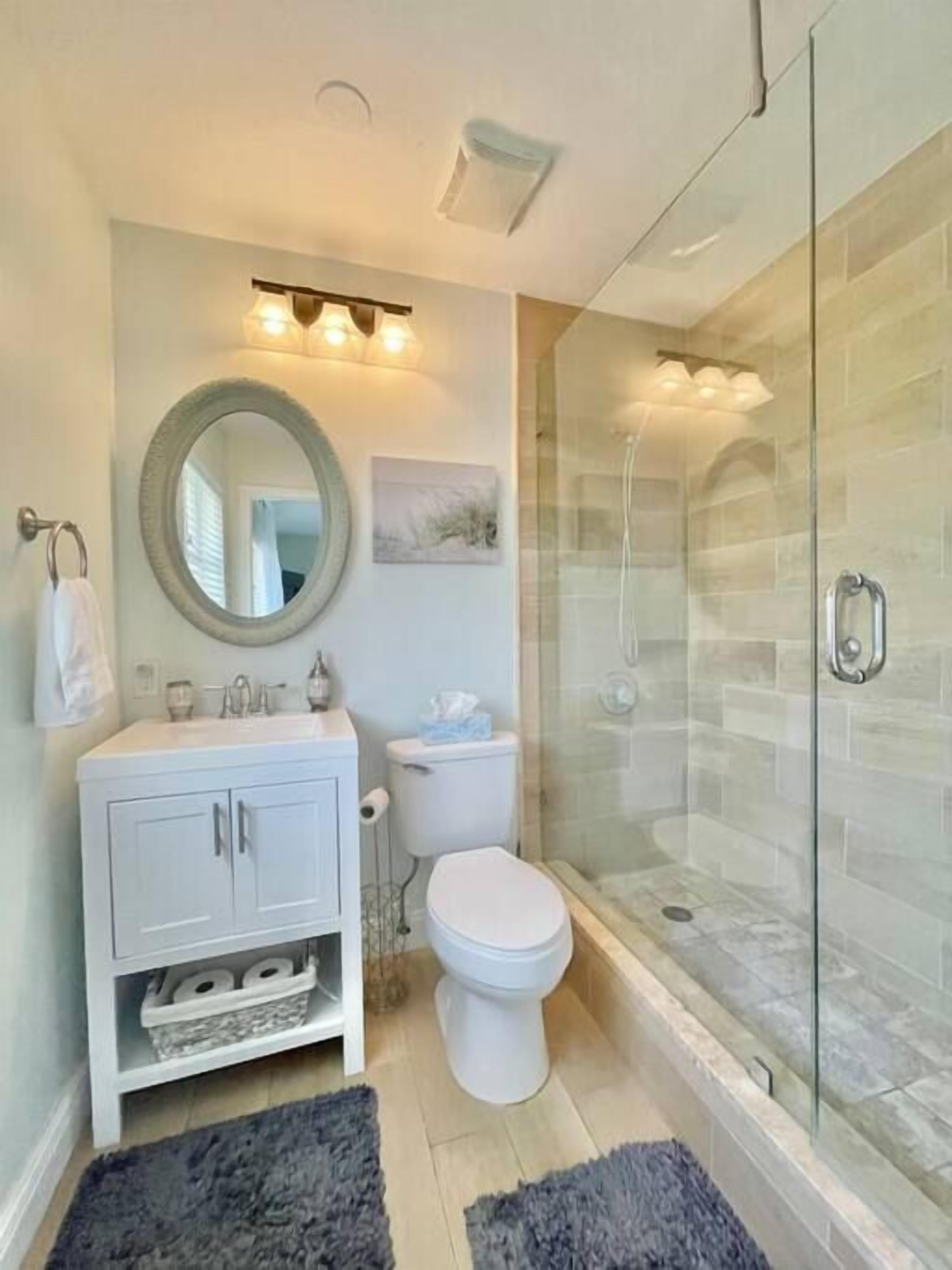 Un baño moderno con ducha de vidrio, tocador blanco con armarios, espejo redondo y un inodoro.