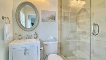 Un baño moderno con ducha de vidrio, tocador blanco con armarios, espejo redondo y un inodoro.