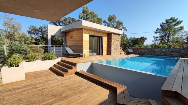 A modern outdoor pool area with a wooden deck, stone walls, and a small building with a glass door.