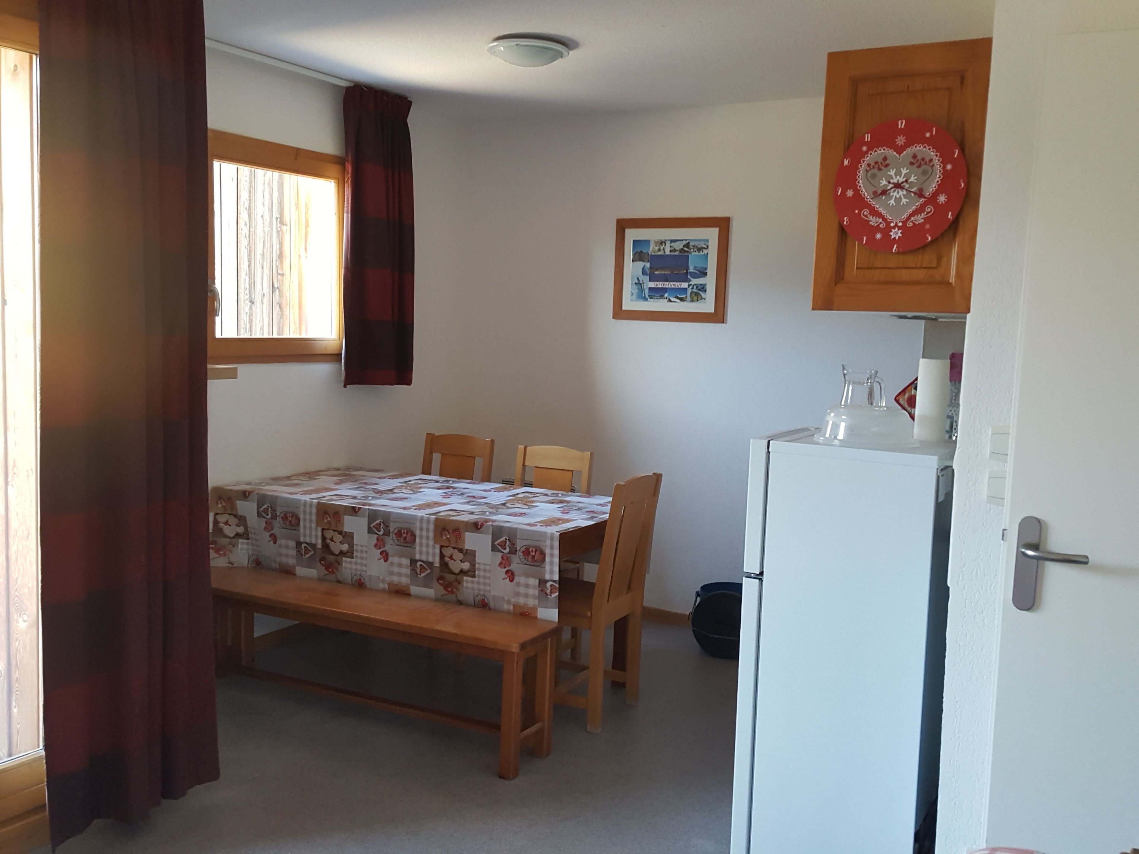 Ett kompakt kök med ett matbord, stolar, ett kylskåp och ett fönster med gardiner.