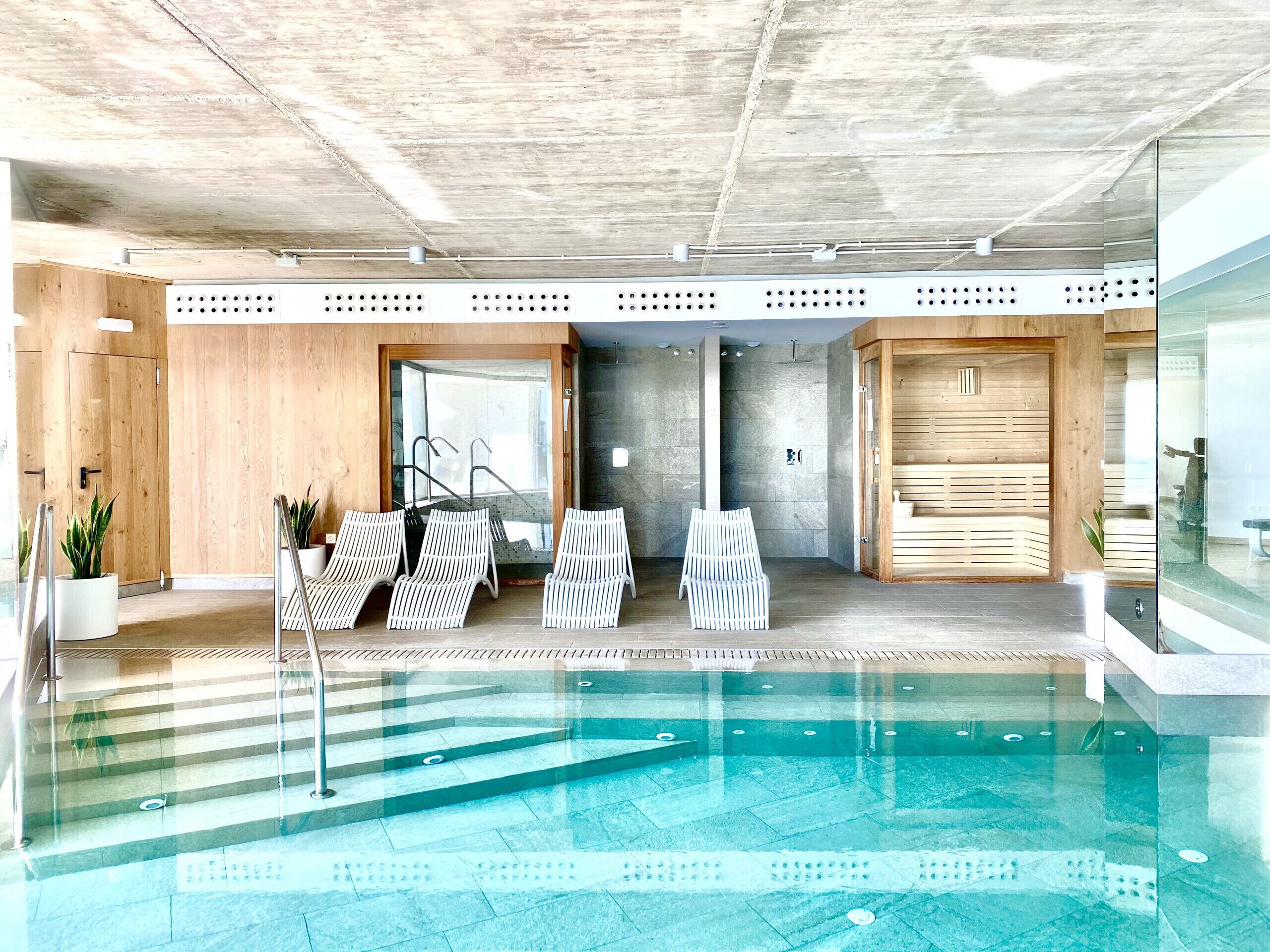 A modern indoor pool area with clear glass walls, a tiled floor, and several lounge chairs.