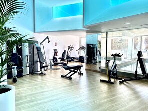 A modern gym with various exercise equipment, large windows, and a wooden floor.