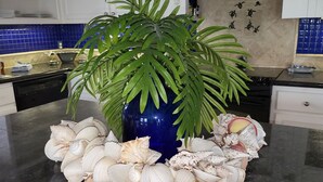 A kitchen counter with a decorative centerpiece featuring a blue vase, a potted plant, and a collection of seashells. A sign reads "WELCOME TO THE BEACH HOUSE."