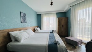 A hotel room with two beds, a wooden wardrobe, a bench, and a large window with sheer curtains.