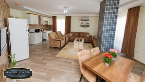 A modern living room with a sofa, coffee table, and dining table. There is a kitchen area with a refrigerator, microwave, and cabinets. The room has wooden flooring and a large window with curtains.