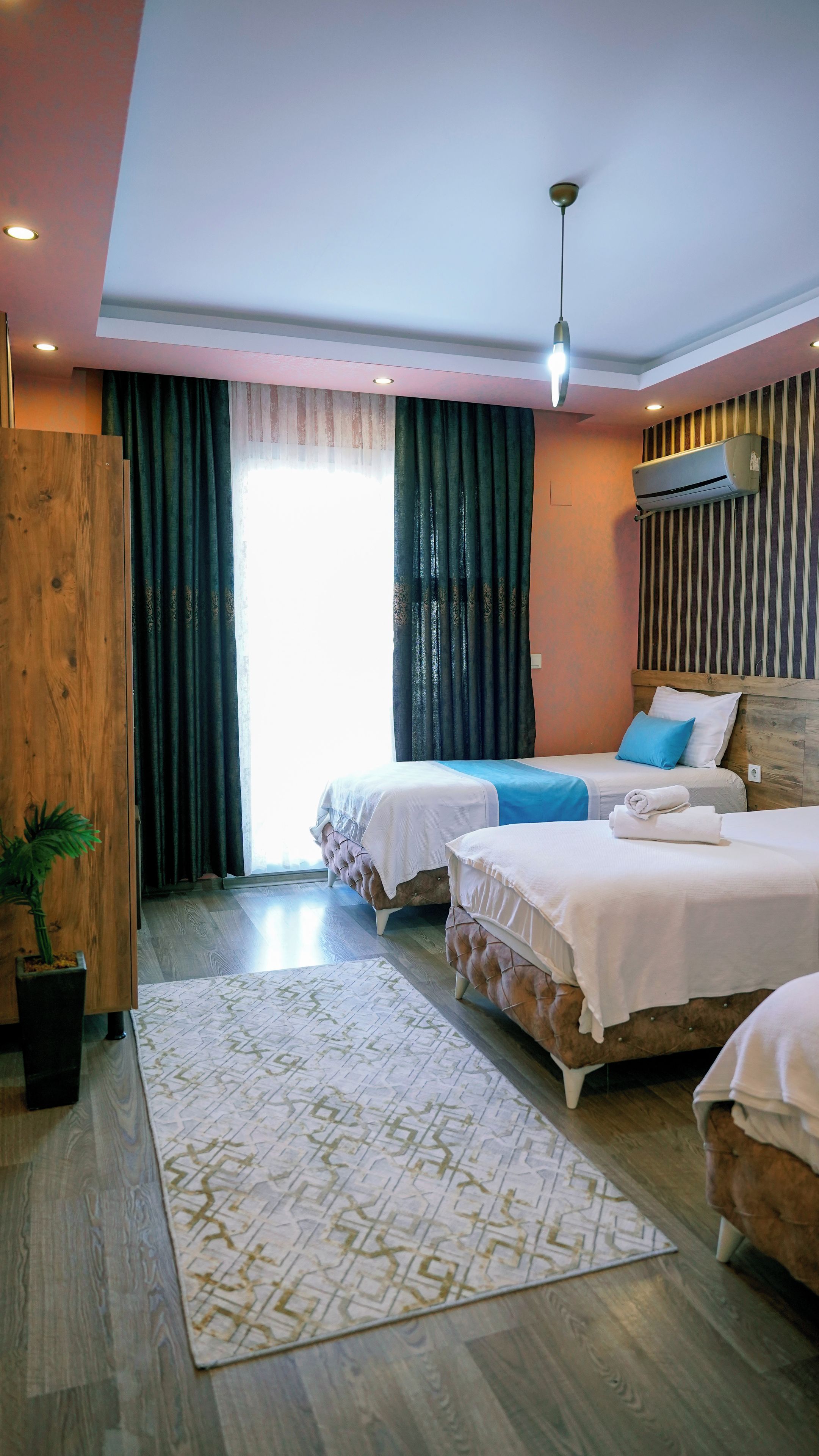 A hotel room with two beds, a wooden wardrobe, a potted plant, and a large window with curtains.